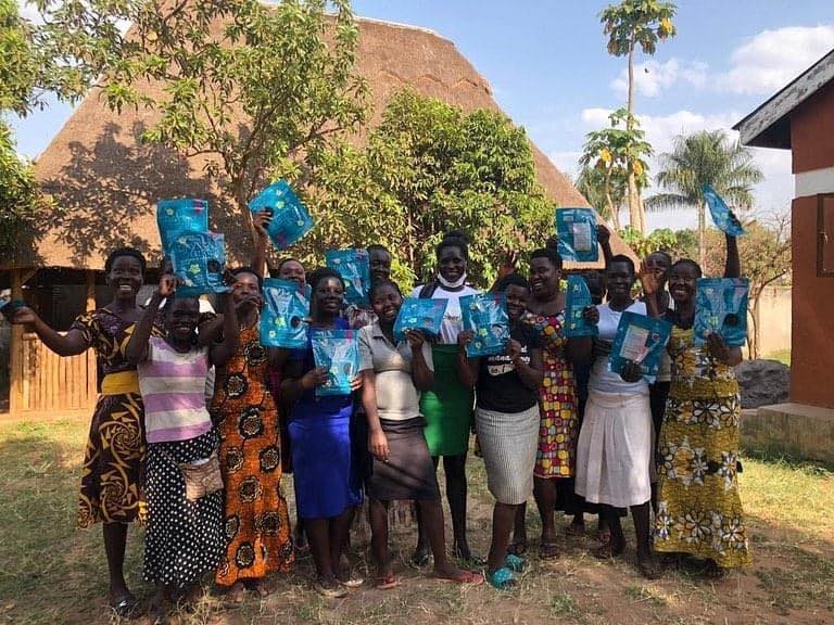 SEW women celebrating together outdoors, holding up blue sanitary packs and smiling at the camera — a joyful moment of community in Lira, Uganda